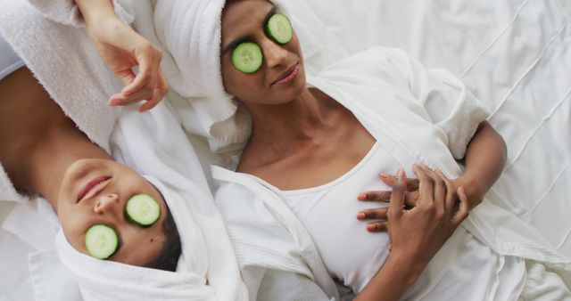 Women in white robes relaxing with cucumber slices over their eyes while lying on a bed, suggesting tranquility and rejuvenation. Ideal for promotions related to skincare, wellness retreats, beauty products, self-care routines, and spa services advertisements.