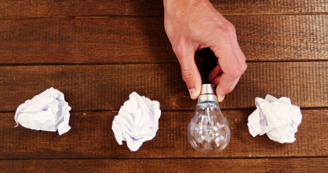 Hand Holding Light Bulb with Crumpled Paper Balls on Wooden Table - Download Free Stock Images Pikwizard.com