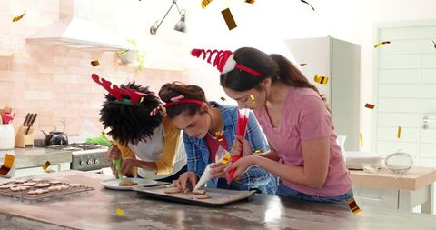 Friends decorating holiday cookies at home kitchen island with piping bags and confetti