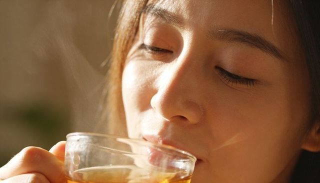 Asian woman sipping steaming tea from clear glass cup in warm golden-hour morning closeup