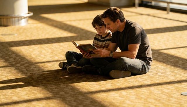 Father reading with son sitting cross-legged in sunlit airport terminal during travel