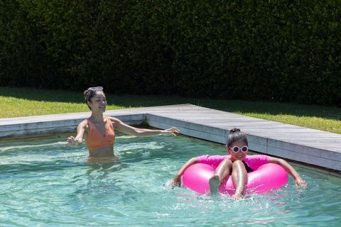 African mother guiding daughter on pink inflatable ring in backyard pool on sunny day
