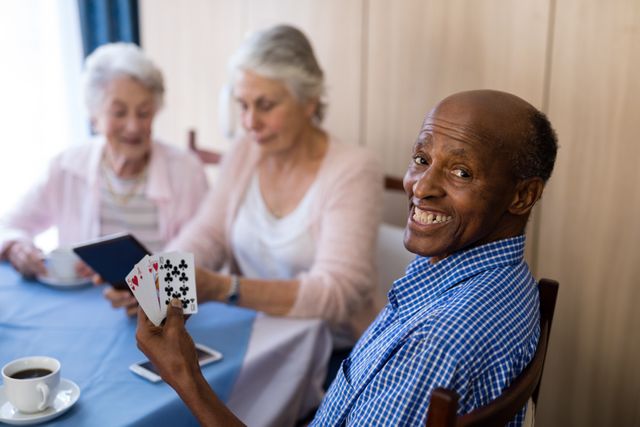 Senior man smiling while playing cards with friends at a table. Ideal for illustrating concepts of leisure, socializing, and community among elderly people. Can be used in advertisements for retirement homes, social clubs, or healthcare services aimed at seniors.
