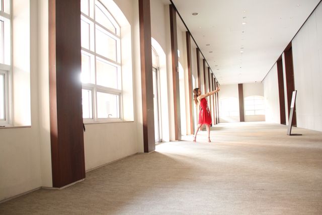 Woman in red dress dancing gracefully in spacious, sunlit empty hallway with large windows, modern architecture. Ideal for concepts of freedom, creativity, movement, dance performance, tranquility. Perfect for use in lifestyle, architectural, dance or artistic blog posts, websites, promotional materials, inspiration for design ideas.