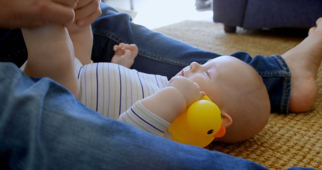 Baby lying on rug, holding yellow rubber duck, with parent nearby. Ideal for concepts of playtime, parenting, early childhood, family bonding, baby toys, and relaxed household moments.