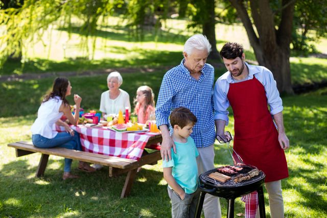 Multi-generational family enjoying a barbeque in a park. Grandfather, father, and son are grilling meat while other family members sit at a picnic table in the background. Ideal for use in advertisements, family-oriented content, and promotional materials for outdoor activities and gatherings.