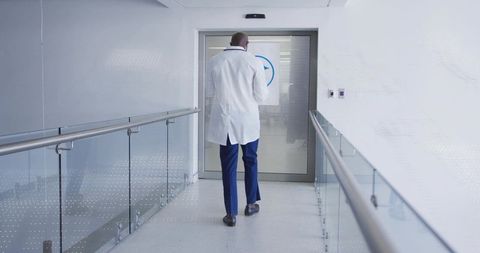 Walking African American Doctor in White Lab Coat Down Modern Glass Medical Corridor