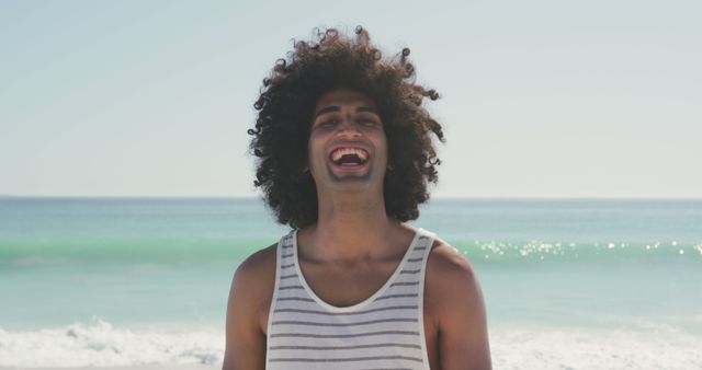 Image shows a man with curly hair and a striped tank top, smiling joyfully at a beach. The ocean waves are visible in the background, along with a clear, sunny sky. Perfect for use in travel and tourism promotions, summer advertisements, lifestyle blogs, and social media campaigns highlighting positivity and leisure.