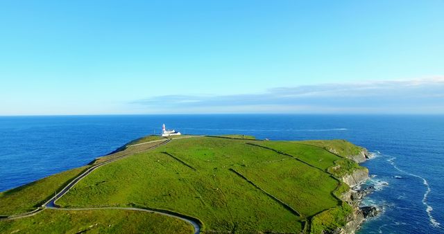 Aerial view of a lighthouse perched on a lush green headland overlooking the ocean, with winding paths leading up to it. The clear blue sky and calm sea create a serene and picturesque landscape ideal for postcards or tranquil backgrounds.