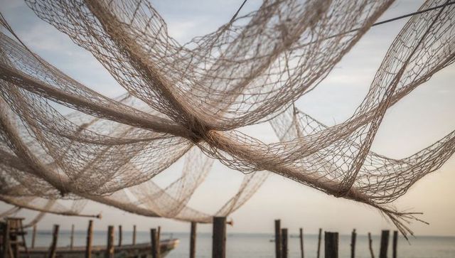 Draping handwoven fishing nets over weathered pier, calm coastal harbor at dawn