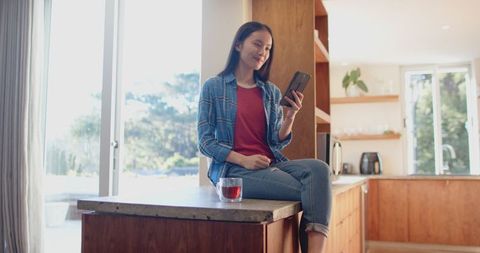 Young Asian Woman Relaxing in Modern Kitchen with Smartphone