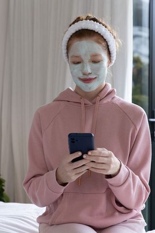 Young woman wearing face mask and pink hoodie using smartphone during self-care routine