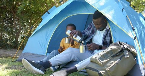 African american father pouring coffee for son at blue tent during camping bonding