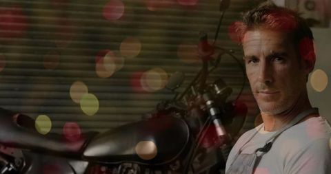 Middle-aged mechanic inspecting custom motorcycle in moody garage with bokeh lights