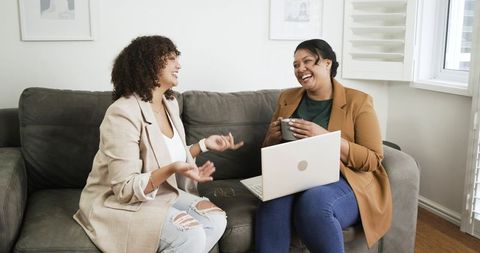 Multiracial women laughing and chatting on sofa with laptop and coffee mug, casual teamwork