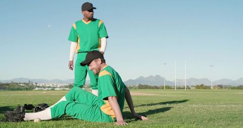 Two male baseball teammates relaxing on grassy field in green uniforms with glove and ball