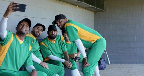 Diverse baseball teammates taking dugout selfie wearing green yellow jerseys