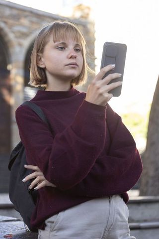 Young female student checking smartphone on campus steps wearing burgundy sweater, backpack