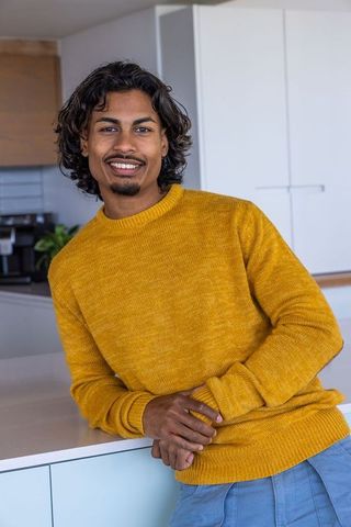 Indian man leaning on kitchen counter wearing mustard sweater smiling, modern home casual