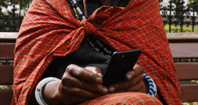 Person wearing traditional African attire, specifically featuring orange plaid cloak and beaded accessories, using smartphone while sitting on bench outdoors. Captures juxtaposition of cultural heritage with modern technology. Useful for illustrating themes of cultural integration, modern technology adoption, African lifestyle, and traditional attire.