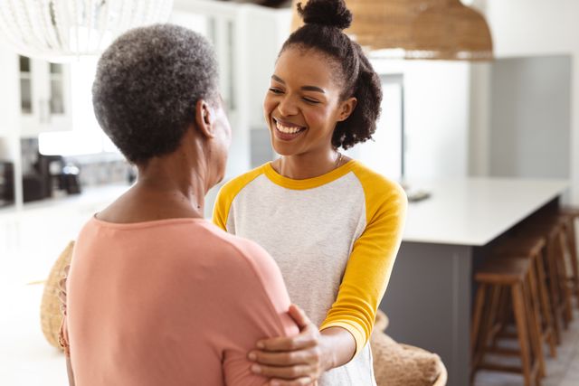This image captures a heartwarming moment between a young African American daughter and her senior mother at home. The daughter is holding her mother's shoulders and smiling warmly, showcasing a strong bond and mutual affection. This image is perfect for use in family-oriented content, advertisements promoting family values, retirement living, healthcare, and lifestyle blogs focusing on intergenerational relationships and happiness.