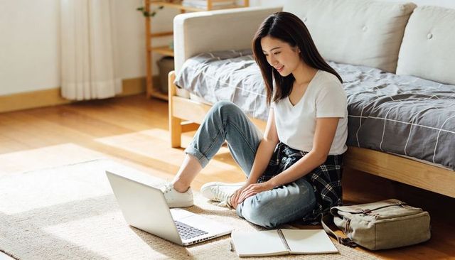 Young asian woman studying at home on laptop sitting on floor with notebook and backpack