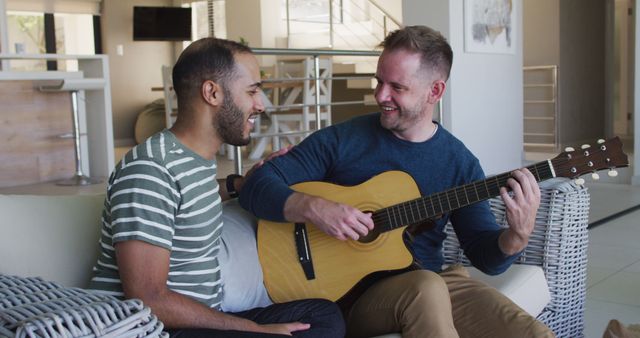 Multi ethnic gay male couple sitting on couch one playing guitar. enjoying staying at home in self isolation in quarantine lockdown.