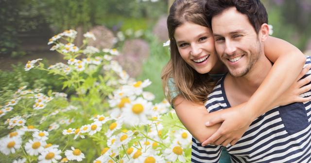 This image shows a joyful couple embracing in a beautiful flower garden filled with daisies. The woman is hugging the man from behind, both smiling and enjoying the outdoors. Perfect for use in advertisements, social media posts, or articles related to love, relationships, nature, and happiness.