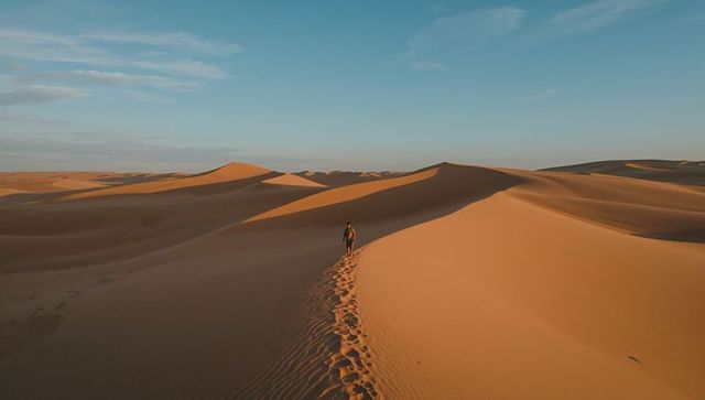 Walking solo traveler crossing vast golden sand dunes at golden hour, desert solitude