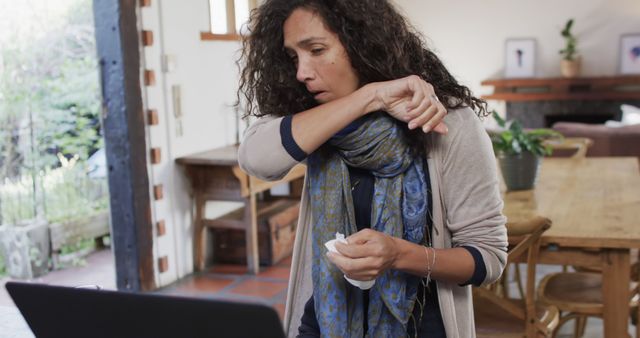 This stock image features a woman coughing while working at a home office desk. She is using a laptop and holding a tissue, suggesting she might be unwell. The environment appears casual with minimal decor, including a scarf indicating she might feel cold. This image can be used for topics related to health, remote work, common cold, flu, illness in the workplace, and work-life balance during illness.