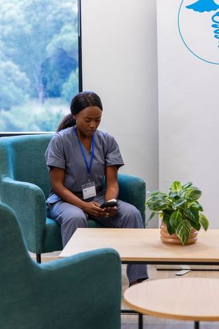 Healthcare worker sitting in clinic lounge checking smartphone and wearing id lanyard