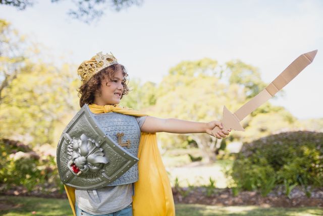 Young boy dressed as a knight, holding a sword and shield, playing in a park. Ideal for themes of childhood imagination, outdoor activities, and playful adventures. Perfect for use in educational materials, children's books, and advertisements promoting creative play.