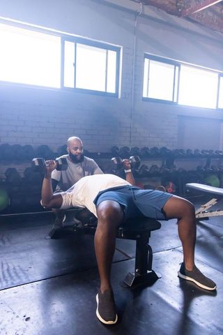 Two african men performing dumbbell bench press with spotter supervising on flat bench