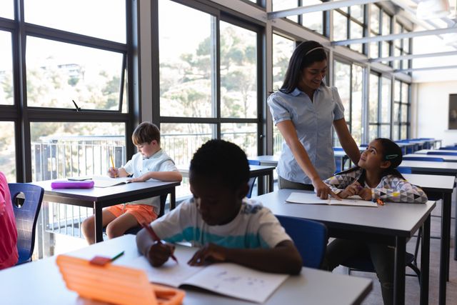 Female teacher assisting students in a bright, modern classroom with large windows. Diverse group of children engaged in learning activities at their desks. Ideal for use in educational materials, school brochures, and articles about teaching methods and classroom environments.