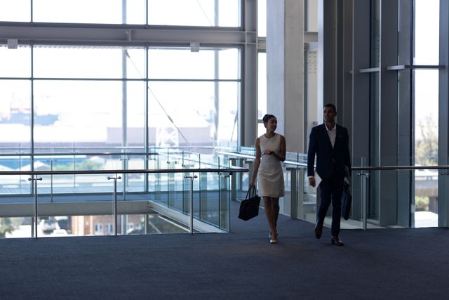 Biracial colleagues walking through a modern office building with large glass windows and natural light. The woman is wearing a dress and carrying a bag, while the man is in a suit, also carrying a bag. This image can be used to depict professional environments, corporate teamwork, business meetings, and modern office settings.