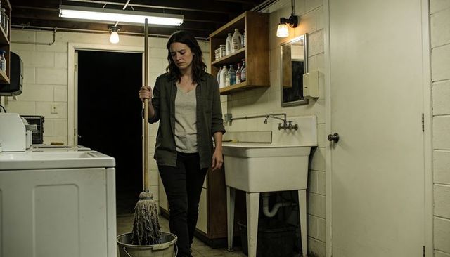 Woman holding mop in dim basement laundry room looking tired and contemplative at night