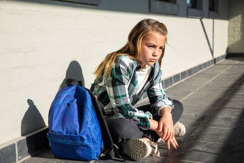 Pensive schoolgirl sitting in sunlit corridor with backpack