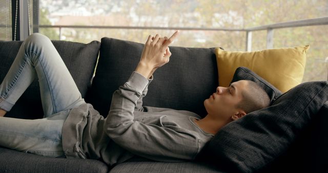 A young adult is lying casually on a dark gray couch, engaging with a smartphone. They are dressed in a gray long-sleeve shirt and light jeans, immersed in the digital experience. The image captures a moment of relaxation and contemporary lifestyle, highlighting elements of modern technology and leisure. Ideal for representing themes like technology usage, relaxation at home, lifestyle trends, and young adult culture in various digital contents and marketing materials.