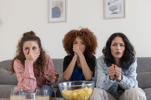 Diverse friends sitting on sofa reacting tensely while watching tv with bowl of chips