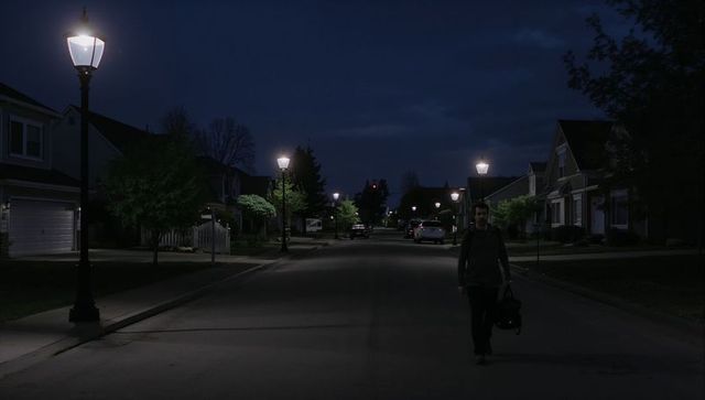 Walking man carrying duffel on dimly lit suburban street under lamp lights at night