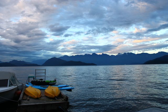 A tranquil sunset scene over a calm lake, with docked boats and colorful kayaks in the foreground. The silhouette of distant mountains enhances the beauty of the scene, adding a sense of peace and adventure. Suitable for use in travel blogs, outdoor adventure promotions, and nature appreciation materials.
