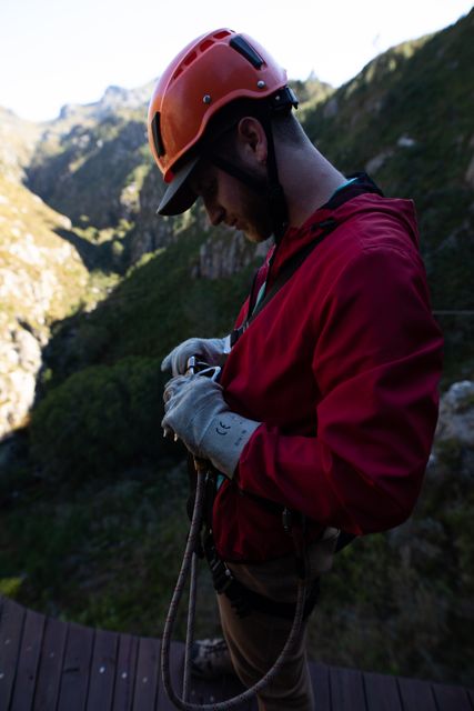 Caucasian man wearing zip lining equipment, standing on a platform in mountainous terrain, preparing his harness. Ideal for use in travel blogs, adventure tourism promotions, outdoor activity advertisements, and safety gear marketing.