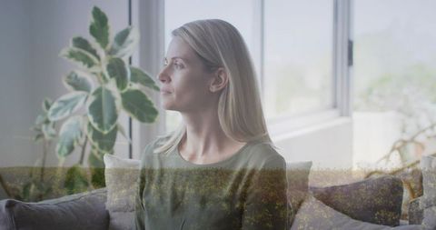 Woman gazing through wide windows on sofa with soft double exposure of wildflowers