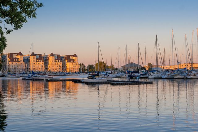 Serene marina setting during sunset with numerous docked sailboats and residential buildings on the waterfront reflecting off the calm water. Ideal for content related to travel, leisure activities, nautical themes, real estate promotions near marinas, and sunset photography.