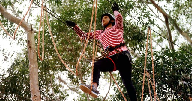 A young girl engages in a high ropes course adventure among the trees, with copy space. She is wearing safety gear and concentrating on her next move, highlighting the importance of confidence and balance in such activities.