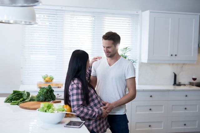 This image shows a happy couple in a modern kitchen, with the man touching the woman's pregnant belly. They are smiling and sharing a moment of joy and anticipation. The kitchen is bright and clean, with fresh vegetables on the counter, suggesting a healthy lifestyle. This image can be used for themes related to family, pregnancy, parenting, home life, and healthy living.