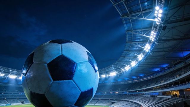 Worn soccer ball sitting on field under stadium floodlights, dramatic blue arena lighting