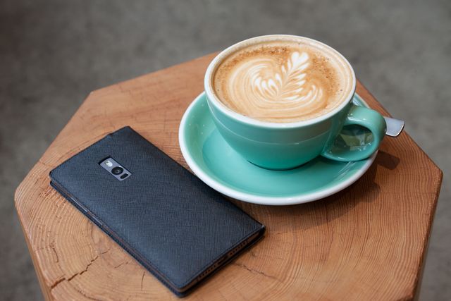 Teal coffee cup with latte art cappuccino next to black smartphone on rustic wooden table. Perfect for illustrations in cafe menus, social media posts about coffee culture, or technology use in leisure settings.