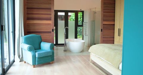 Teal armchair sitting by sliding glass doors with freestanding tub in bright modern bedroom