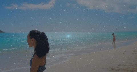 Standing woman wearing dark swimsuit ponytail facing teal sea on sandy beach for summer vacation